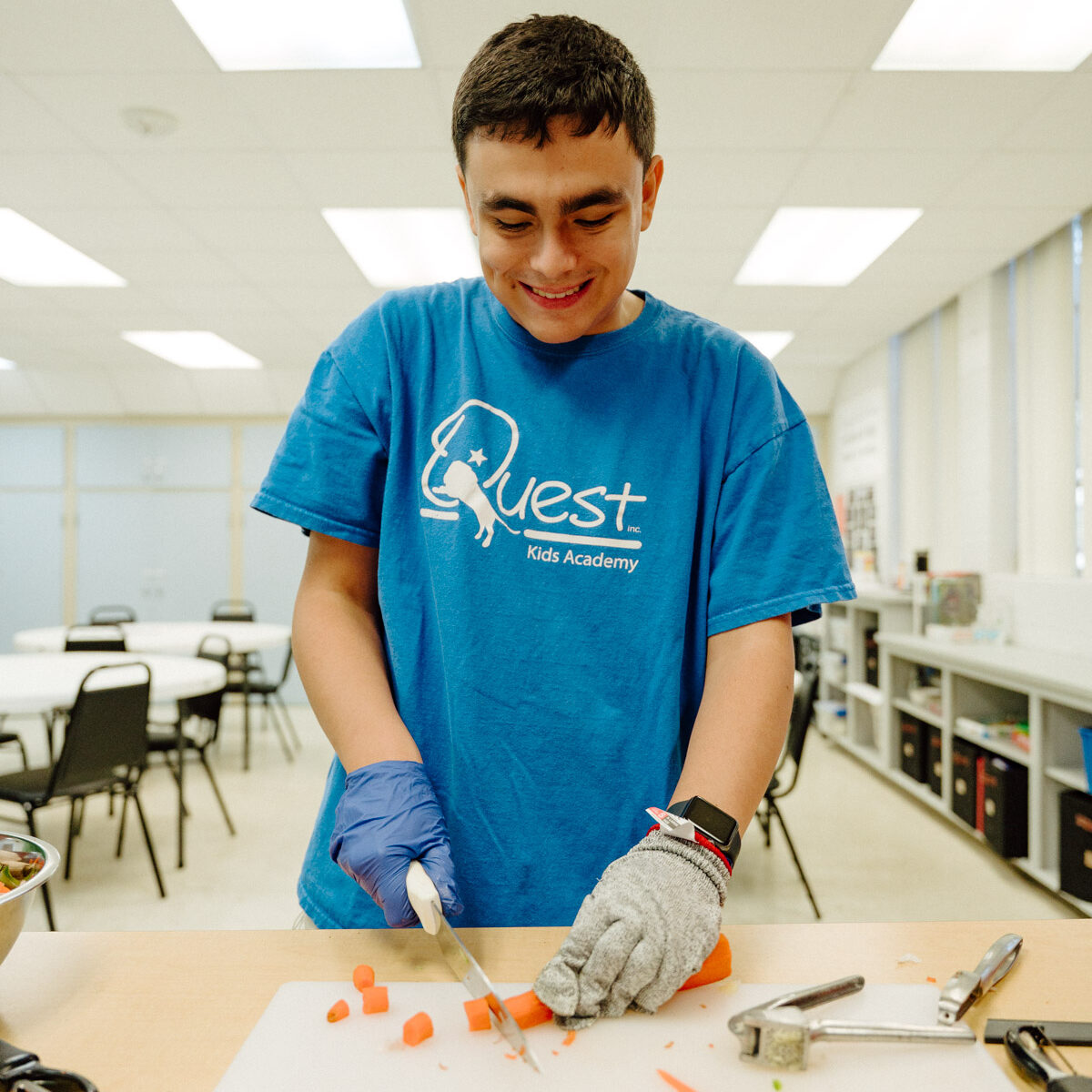 A teenage male student in a blue Quest Kids Academy t-shirt is practicing cooking skills by cutting carrots.