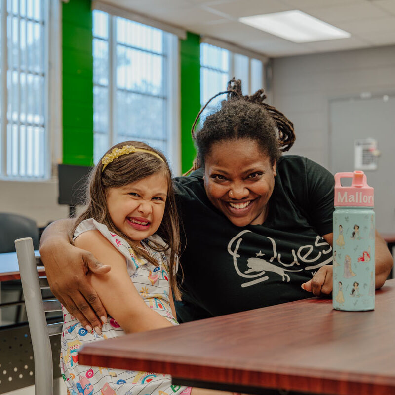 A female Quest Kids Academy teacher has her arm around a female student and they are both smiling at camera.